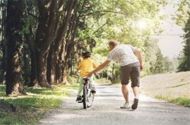 Adult teaching child how to ride a bicycle on a tree-lined pathway on a sunny day.